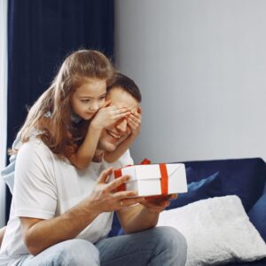 imagem de uma menina tampanto os olhos do pai enquanto segura uma caixa de presente sentado na cama
